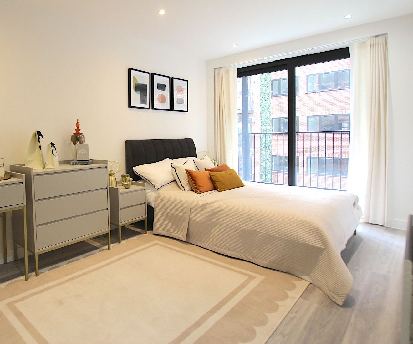 Staged double bedroom with a grey upholstered bed, orange and mustard cushions and floor-to-ceiling windows